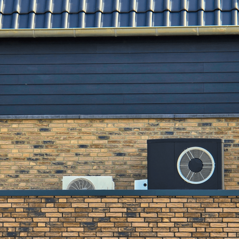 Donkere warmtepompbuitenunit en gedeeltelijk zichtbare lichte buitenunit achter een lage bakstenen muur