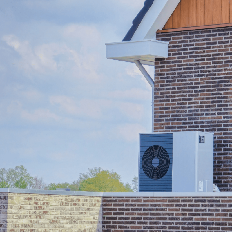 Buitenunit van een warmtepomp op een plat dak naast de bakstenen gevel van een woning
